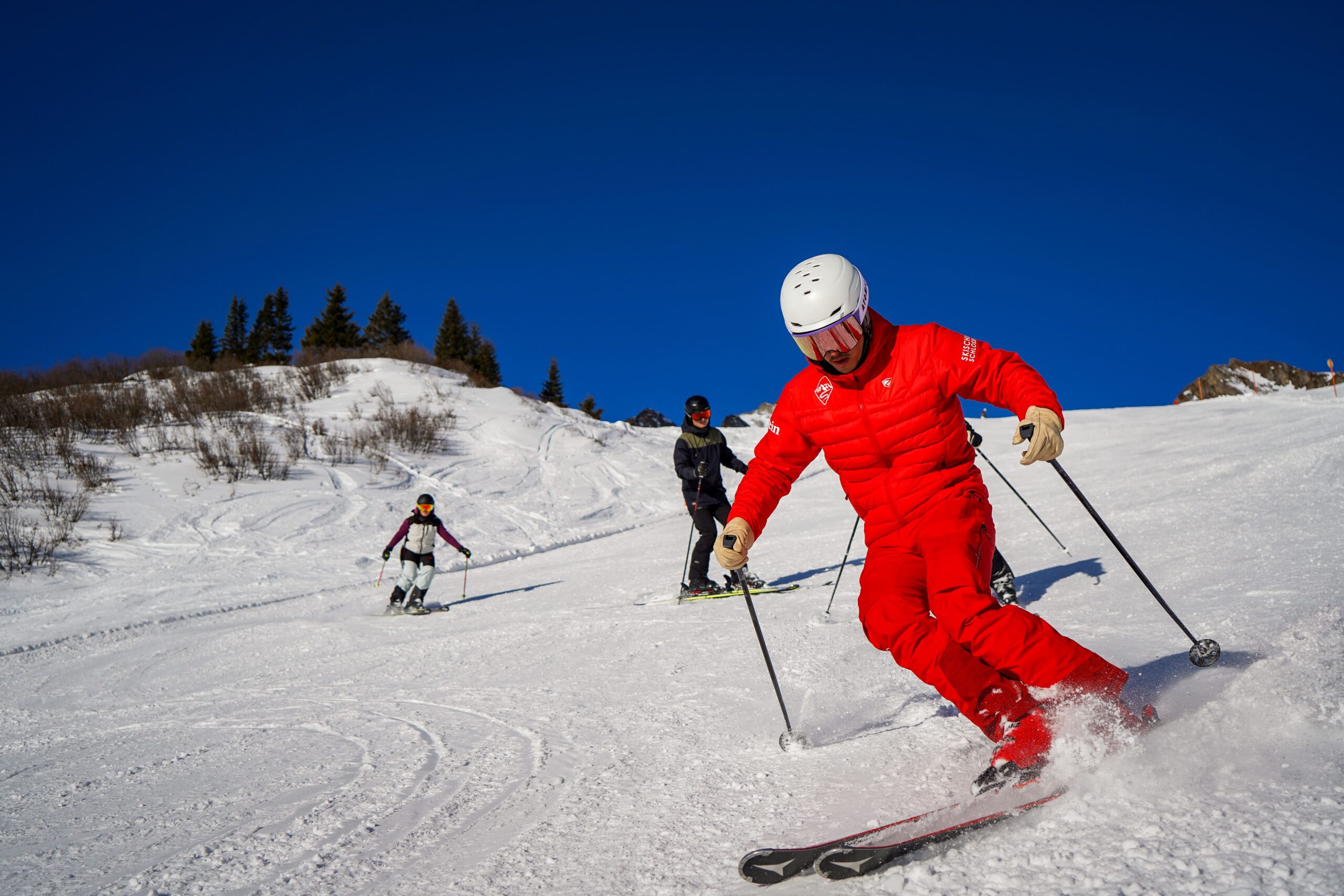 Skilehrer fährt schwungvoll vor einer Skigruppe auf sonniger Piste im Gelände.“